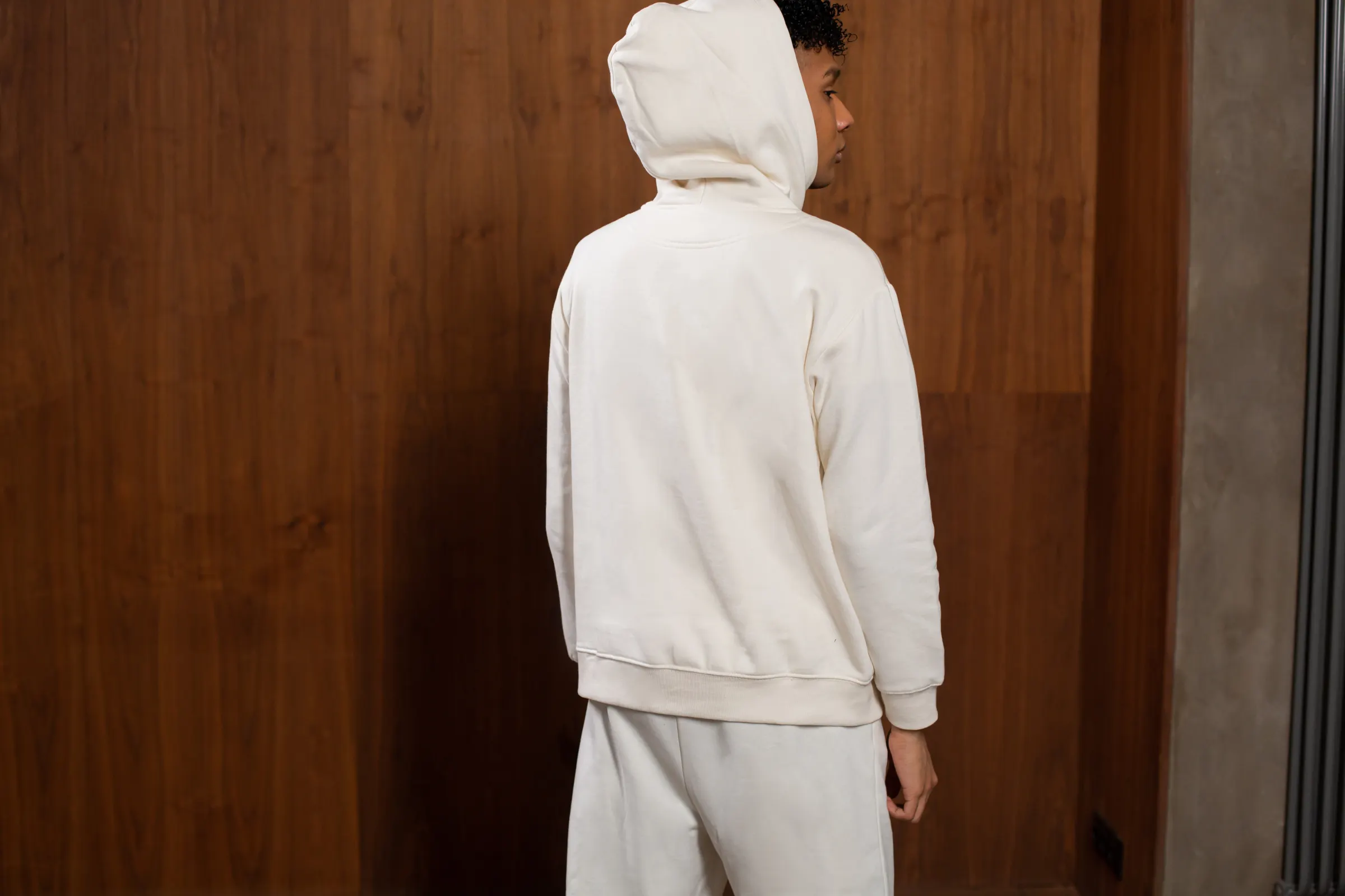 Homem de costas com o capuz do moletom oversized off-white levantado, diante de painel de madeira