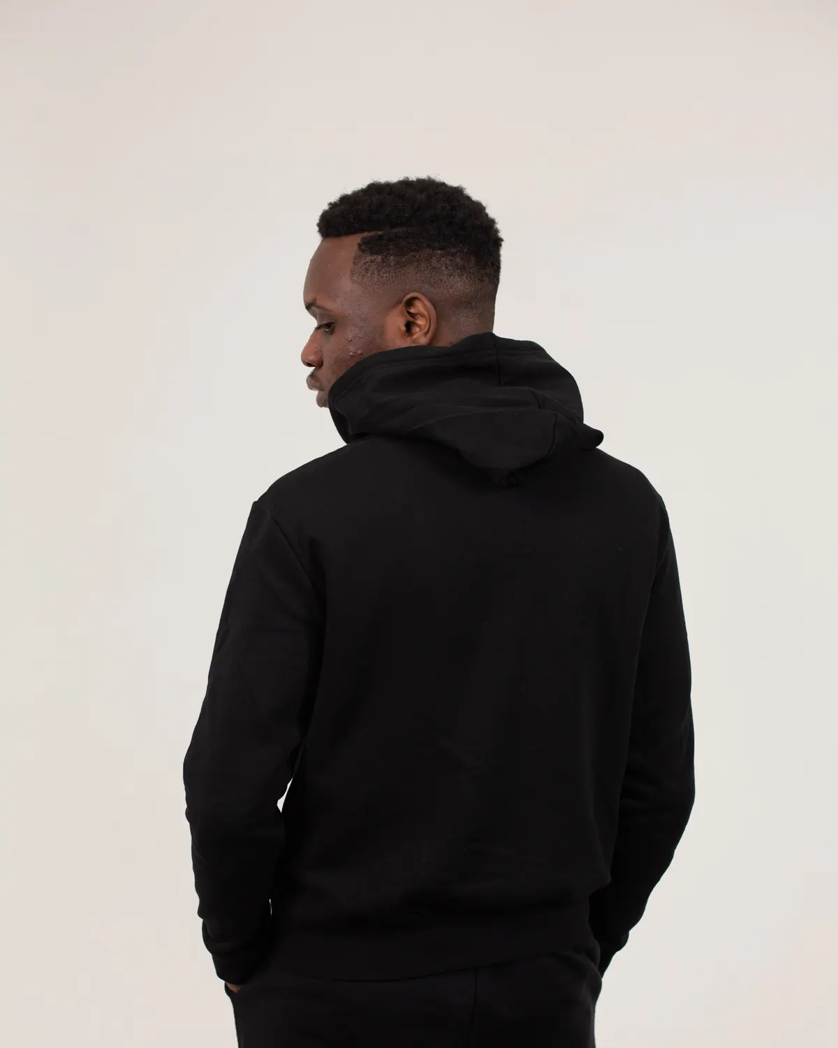 Homem de costas vestindo moletom oversized preto com capuz, em fundo claro de estúdio